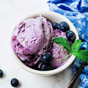 no churn blueberry ice cream in a white bowl garnished with mint sprig on a table with a blue napkin and blueberries scattered around.