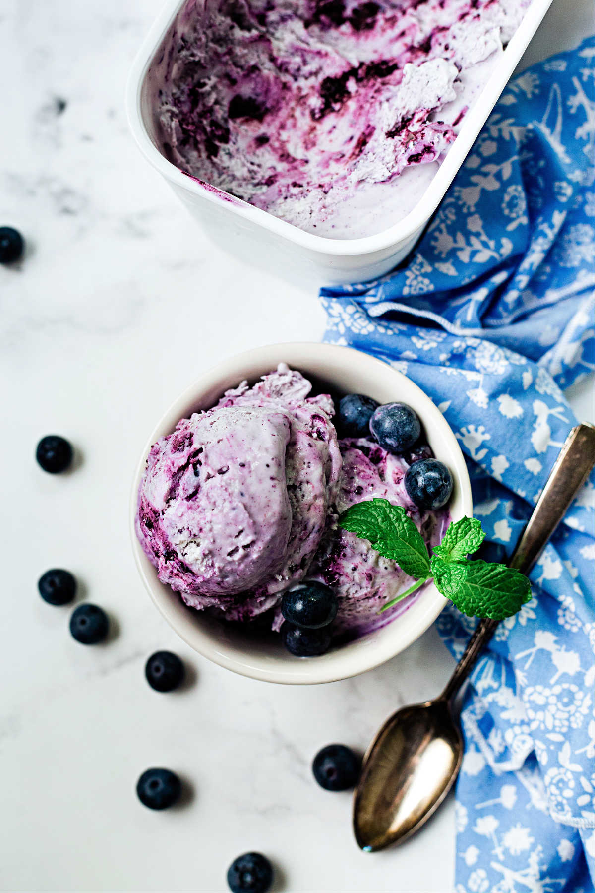 no churn blueberry ice cream in a white bowl garnished with mint sprig on a table with a blue napkin and blueberries scattered around and a container of ice cream.