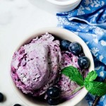no churn blueberry ice cream in a white bowl garnished with mint sprig on a table with a blue napkin and blueberries scattered around.