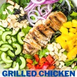 a large salad bowl with salad greens, chopped mango, cucumber, red bell pepper, red onions, and almonds with a sliced grilled chicken breast laying on top.
