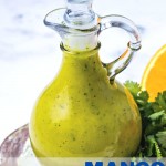 a decorative carafe filled with mango dressing sitting on a table surrounded by cilantro, an orange slice, and a mango.