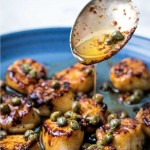 a spoon drizzling brown butter sauce over a plate of seared scallops.