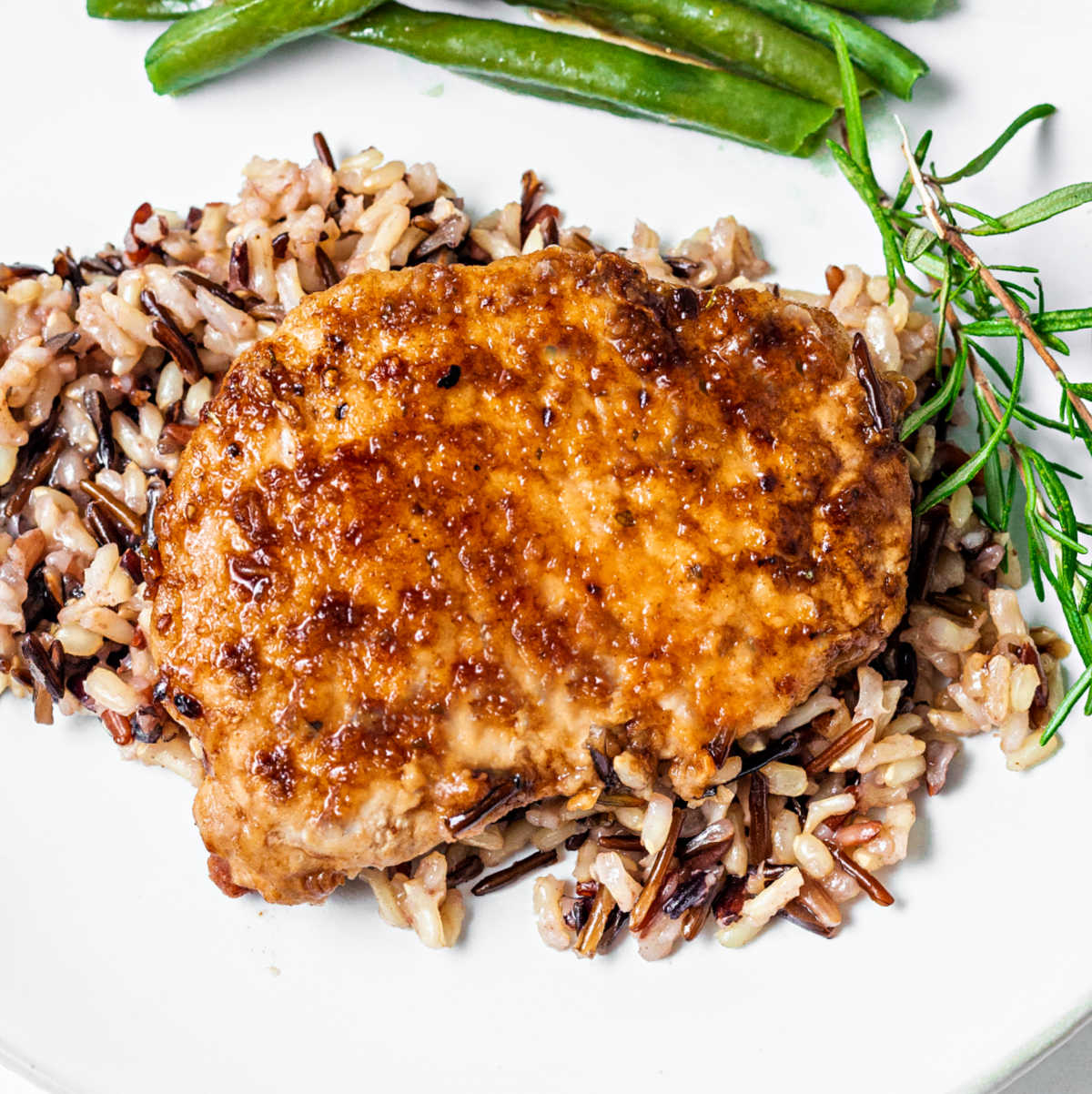 pork chop on a bed of rice on a plate with green beans and a sprig of rosemary on a table.