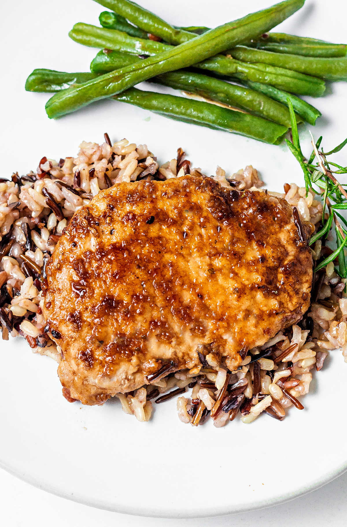 pork chop on a bed of rice on a plate with green beans and a sprig of rosemary on a table.