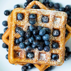 top down view of blueberry waffles stacked on top of each other with fresh blueberries scattered around on a white plate.