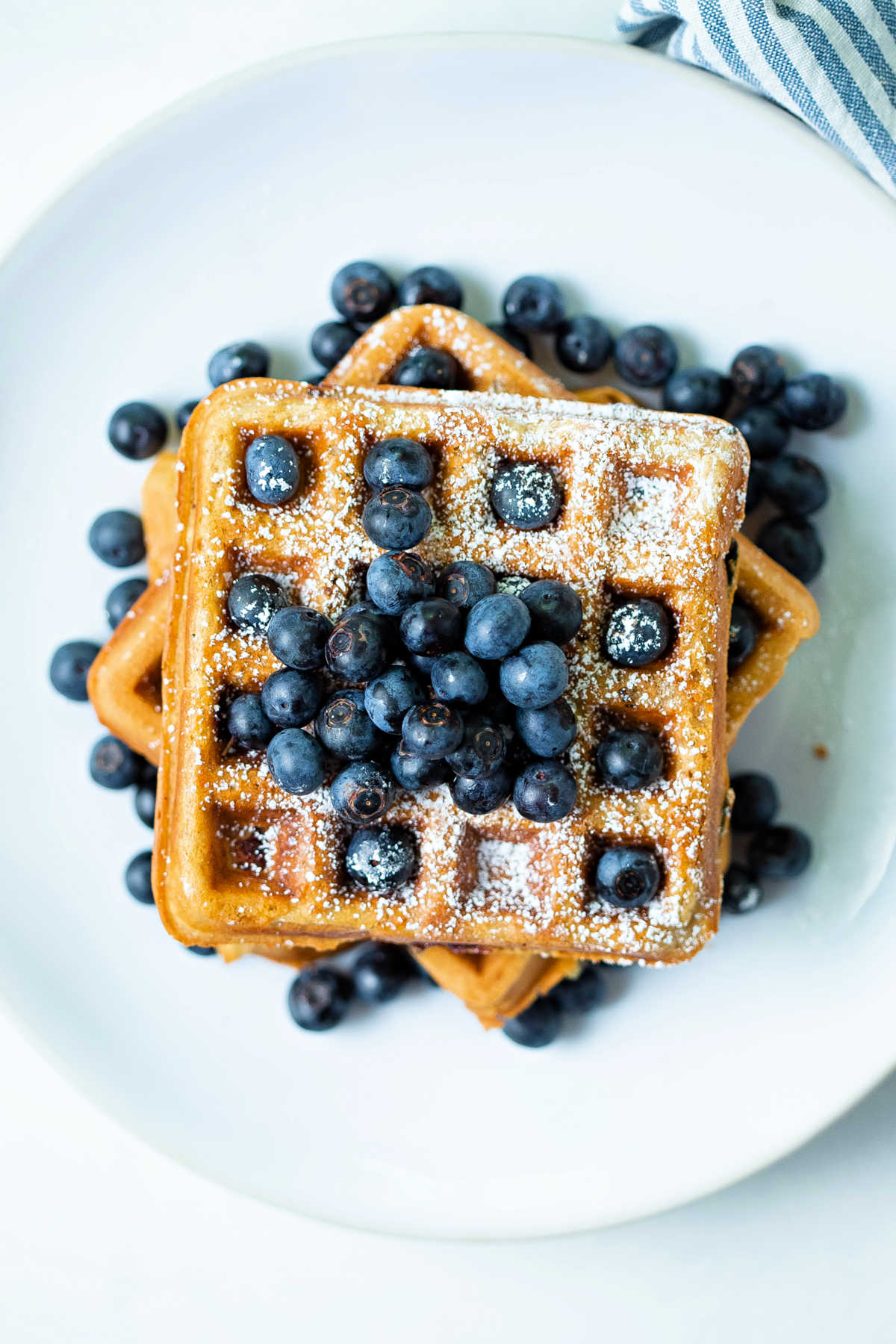 top down view of blueberry waffles stacked on top of each other with fresh blueberries scattered around on a white plate.