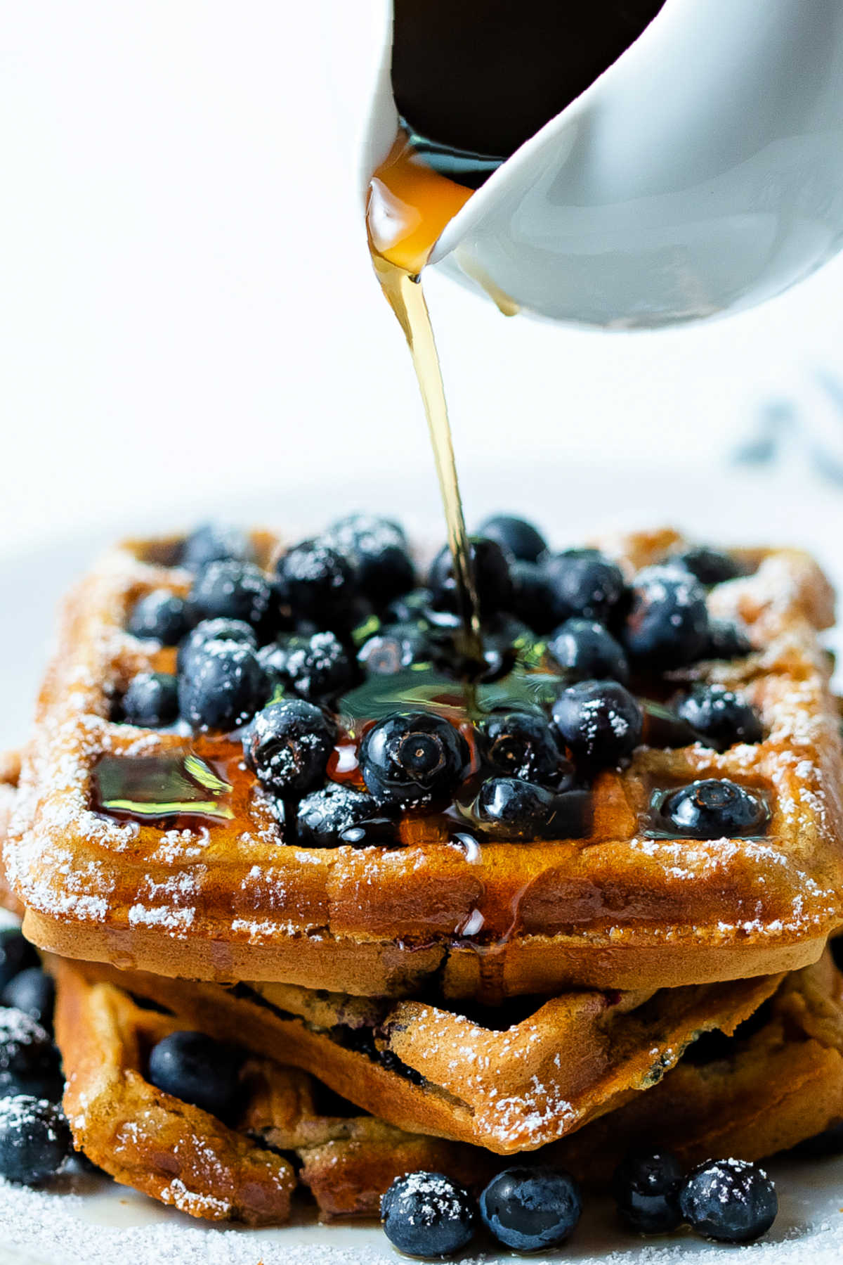 a stack of waffles with a pile of blueberries on top and maple syrup being poured over all.