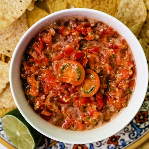 a bowl of cherry tomato salsa on a decorative plate with tortilla chips scattered around.