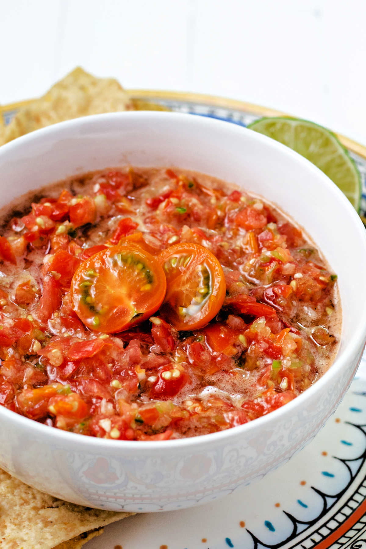 a bowl of cherry tomato salsa on a decorative plate with tortilla chips scattered around.