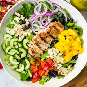 a large salad bowl with salad greens, chopped mango, cucumber, red bell pepper, red onions, and almonds with a sliced grilled chicken breast laying on top.