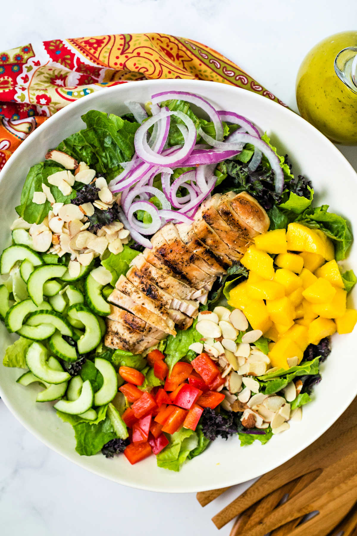 a large salad bowl with salad greens, chopped mango, cucumber, red bell pepper, red onions, and almonds with a sliced grilled chicken breast laying on top.