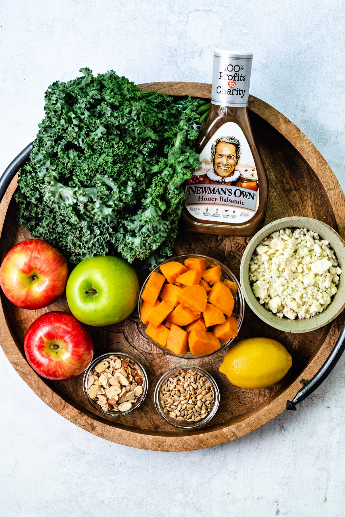 ingredients for harvest chicken salad on a wooden tray: a bunch of curly kale, apples, blue cheese crumbles, balsamic dressing, a lemon, sunflower seeds, a bowl of sweet potato chunks, and almonds.