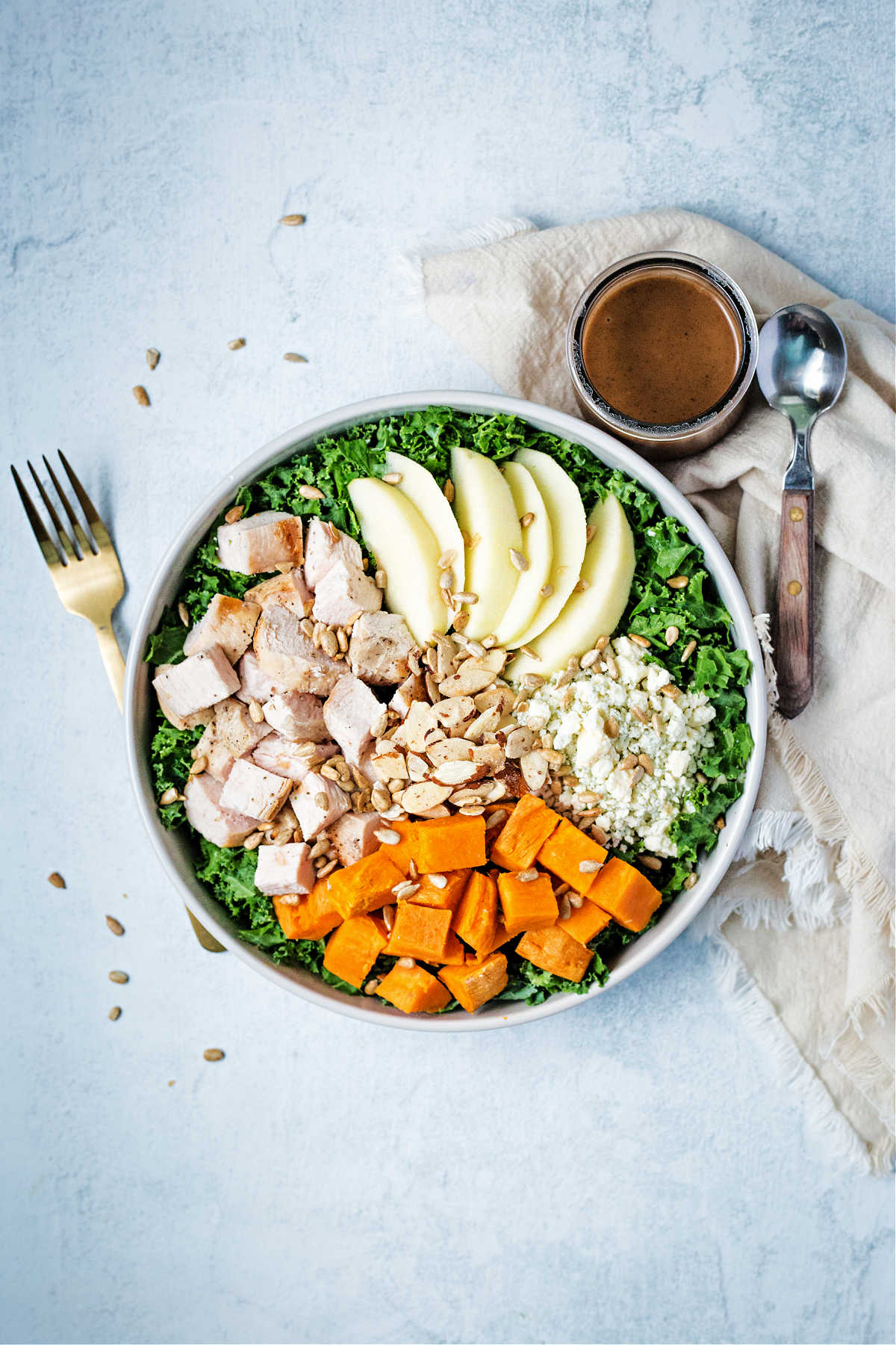 a salad bowl on a table top filled with chopped kale, sweet potatoes, blue cheese, apples, and chicken with a side of balsamic dressing.