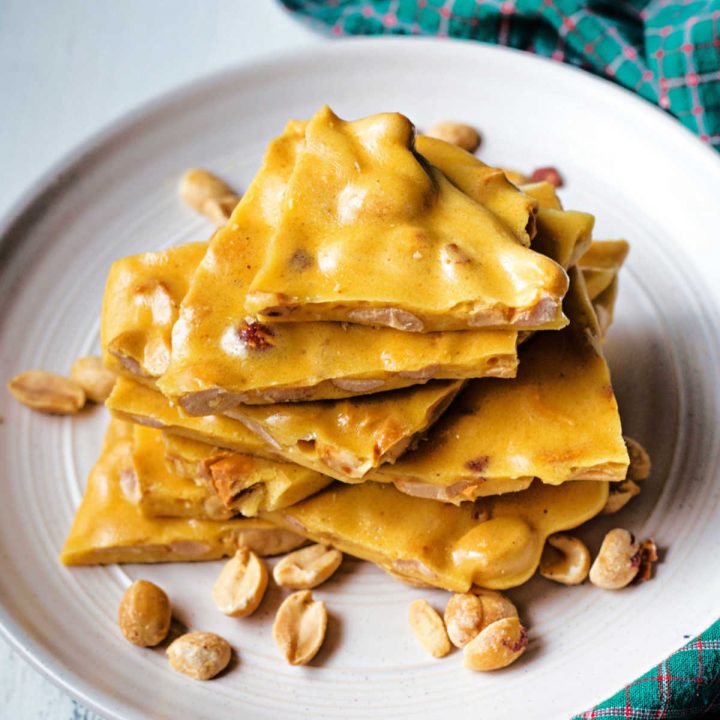 pieces of microwave peanut brittle stacked on a plate with peanuts scattered around.