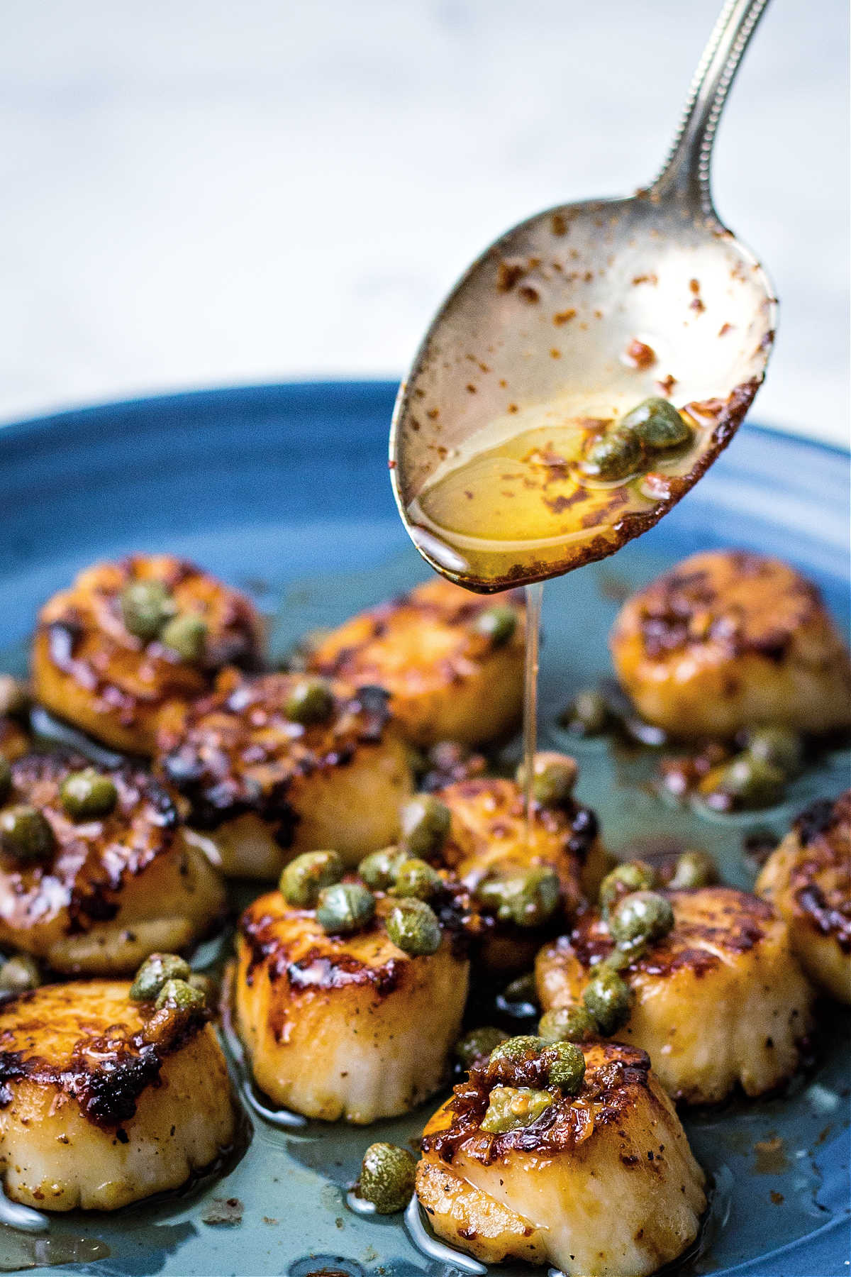 a spoon drizzling brown butter sauce over a plate of seared scallops.