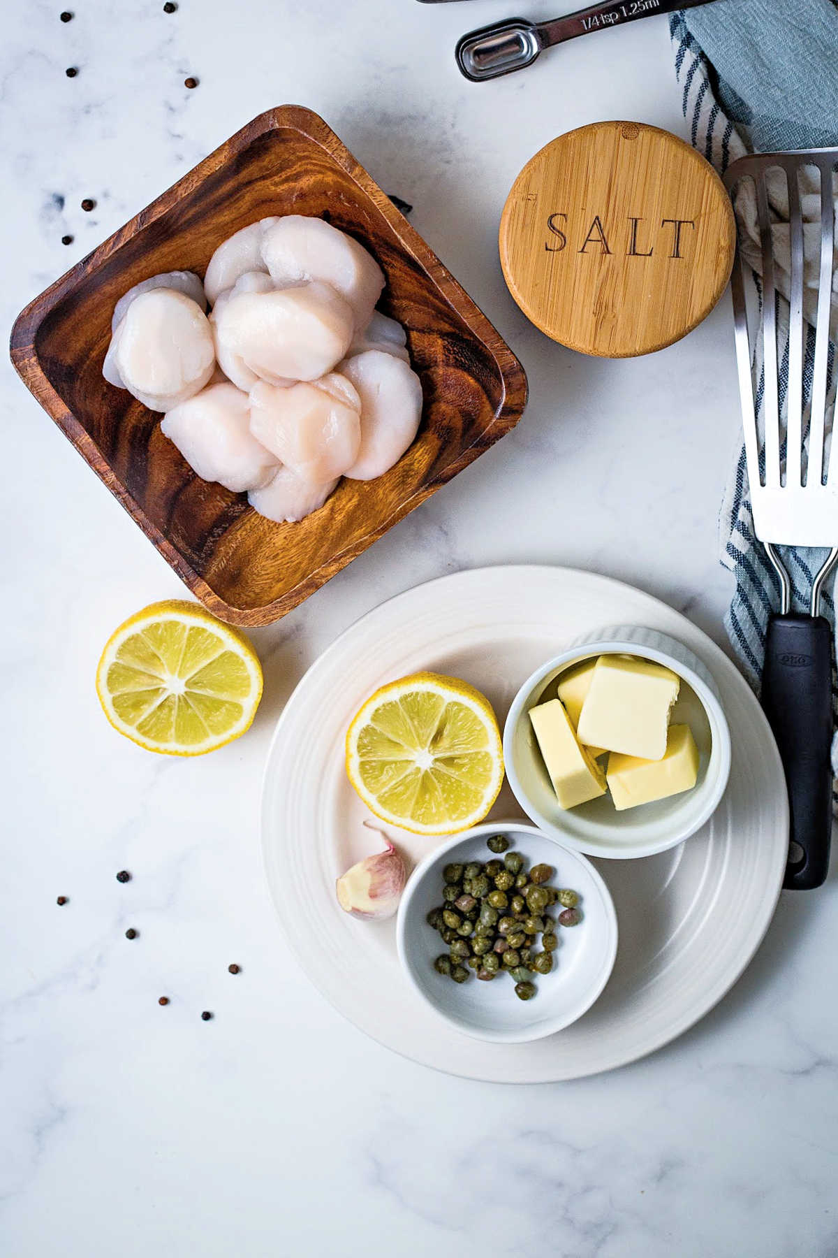 ingredients for making seared scallops on a counter: raw scallops, lemon, capers, butter, and salt.