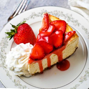 a slice of strawberry cheesecake on a china plate with a mound of whipped cream on the side.