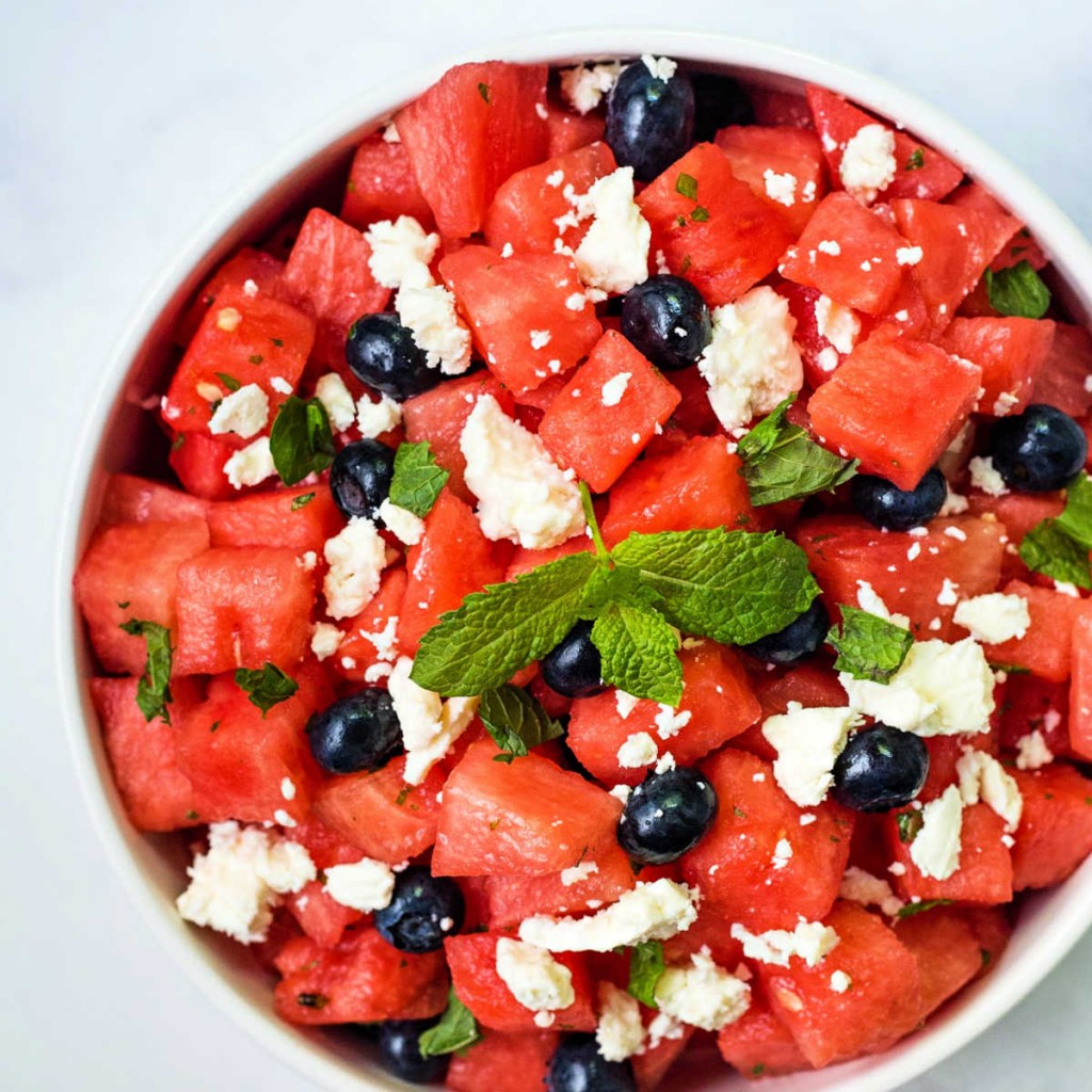cubes of watermelon, blueberries, crumbled feta cheese, and fresh mint in a white bowl on a table.