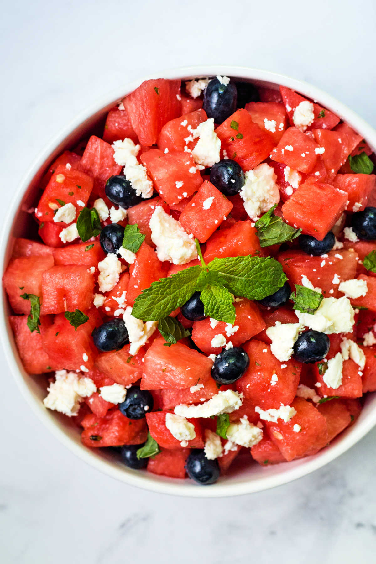 cubes of watermelon, blueberries, crumbled feta cheese, and fresh mint in a white bowl on a table.