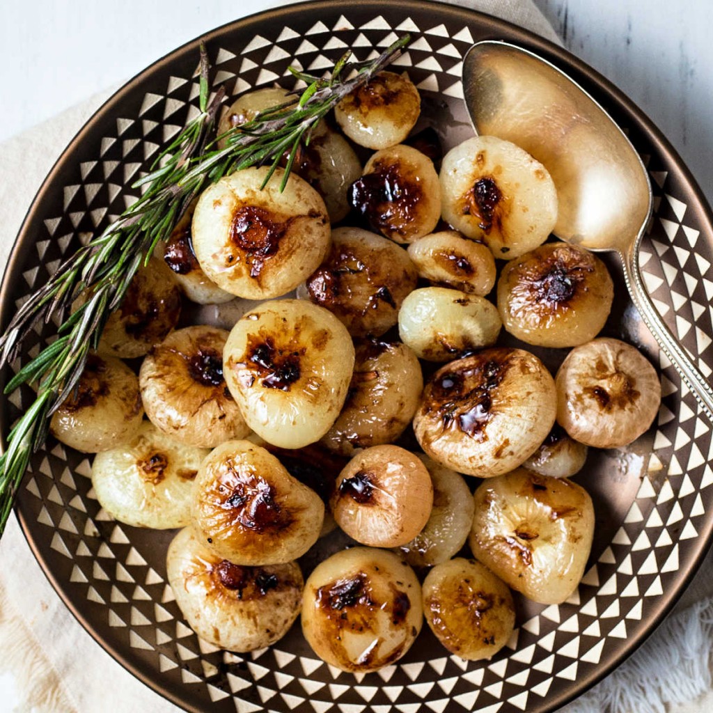 a copper bowl of balsamic glazed cipollini onions garnished with rosemary with a silver spoon to the side.