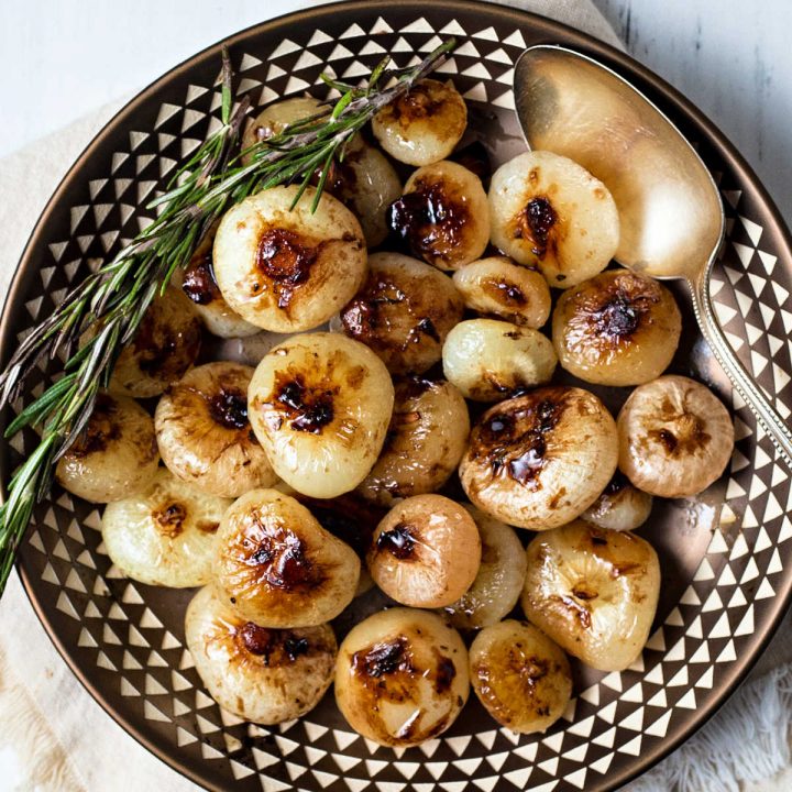 a copper bowl of balsamic glazed cipollini onions garnished with rosemary with a silver spoon to the side.