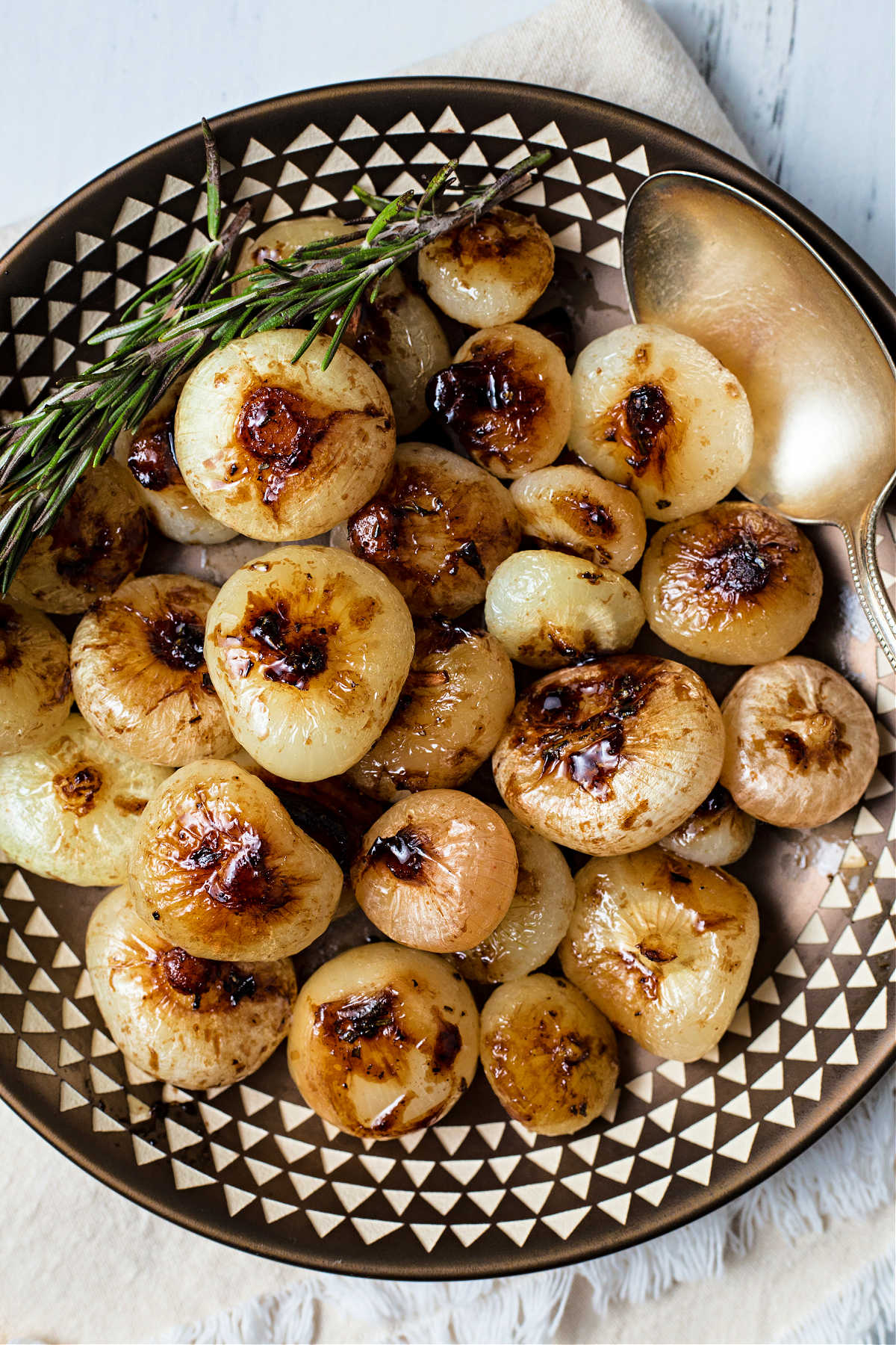 a copper bowl of balsamic glazed cipollini onions garnished with rosemary with a silver spoon to the side.