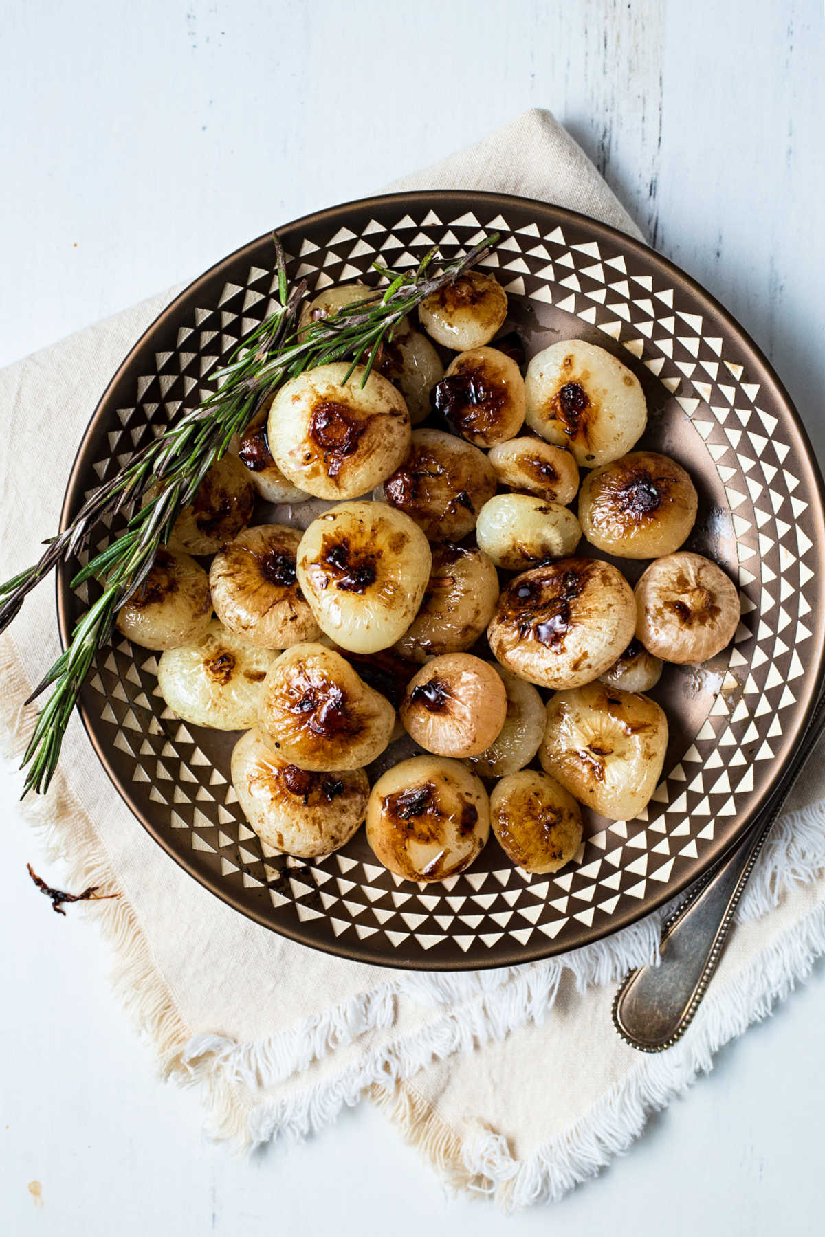 a copper bowl of balsamic glazed cipollini onions garnished with rosemary sitting on a linen napkin.