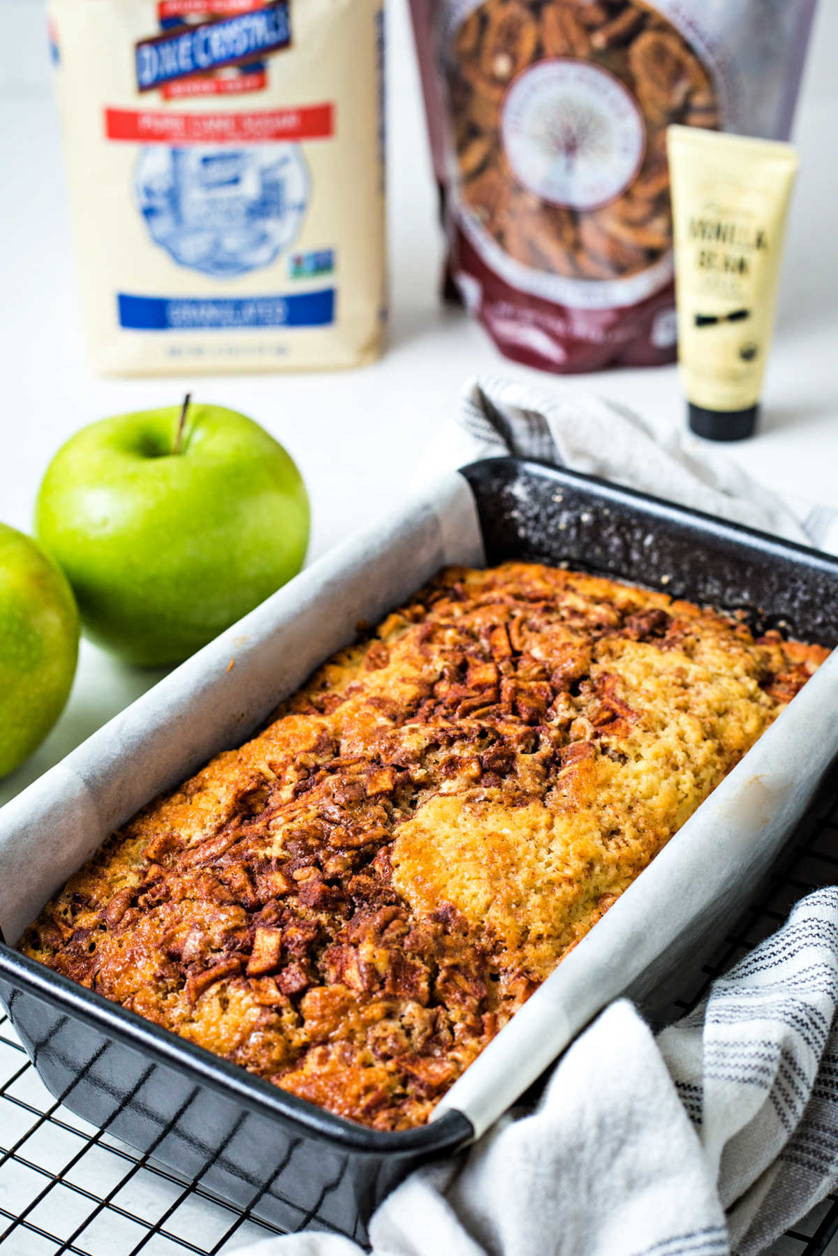 apple cinnamon bread in a loaf pan cooling on a wire rack with apples in the background.