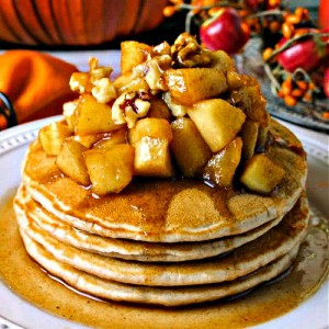 a stack of cinnamon pancakes topped with apple compote and walnuts on a white plate with autumn decor in the background.