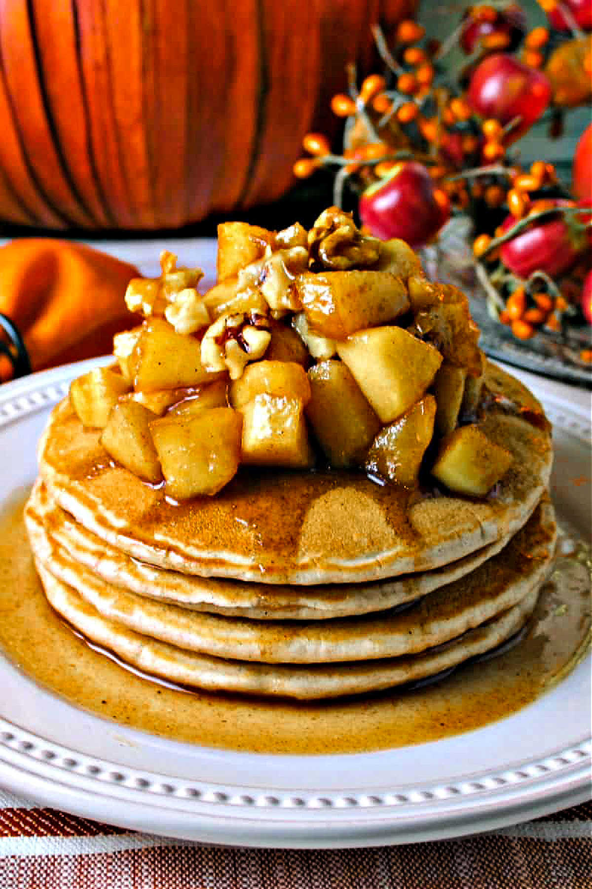 a stack of cinnamon pancakes topped with apple compote and walnuts on a white plate with autumn decor in the background.