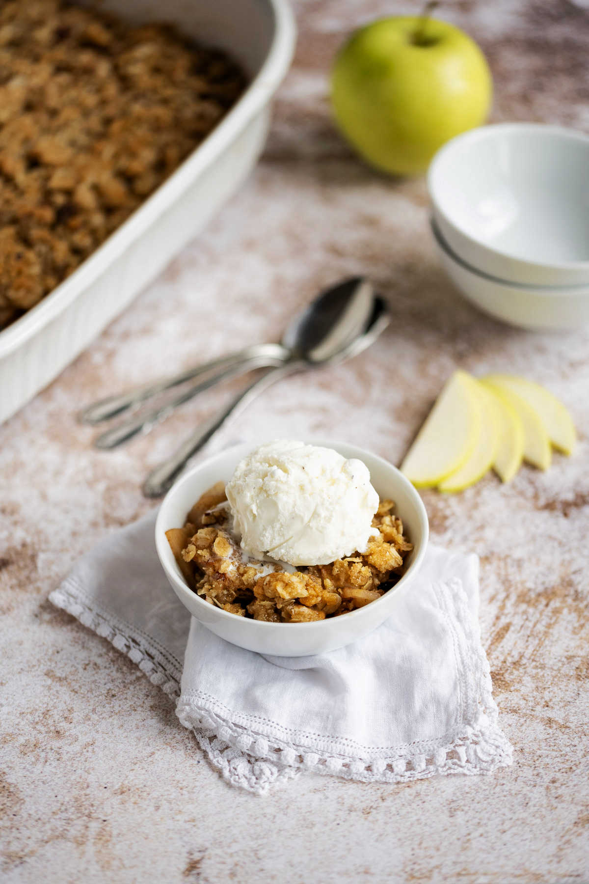 apple crisp in a white bowl with a scoop of vanilla ice cream on top sitting on a counter with apple and baking dish in the background.