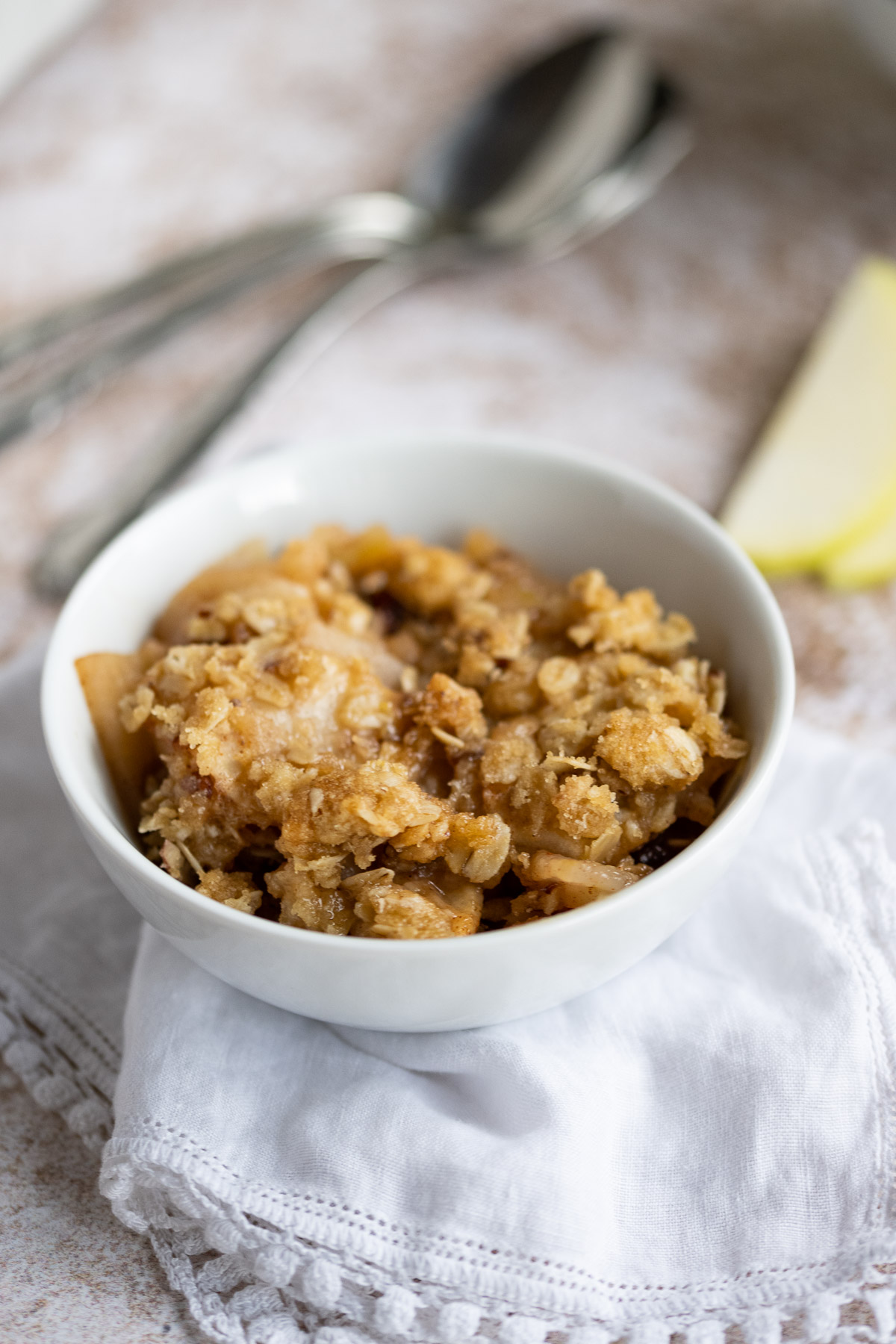 a bowl of apple crisp sitting on a white napkin with spoons in the background.
