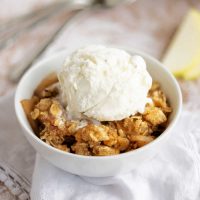 apple crisp in a white bowl with a scoop of vanilla ice cream on top sitting on a counter.