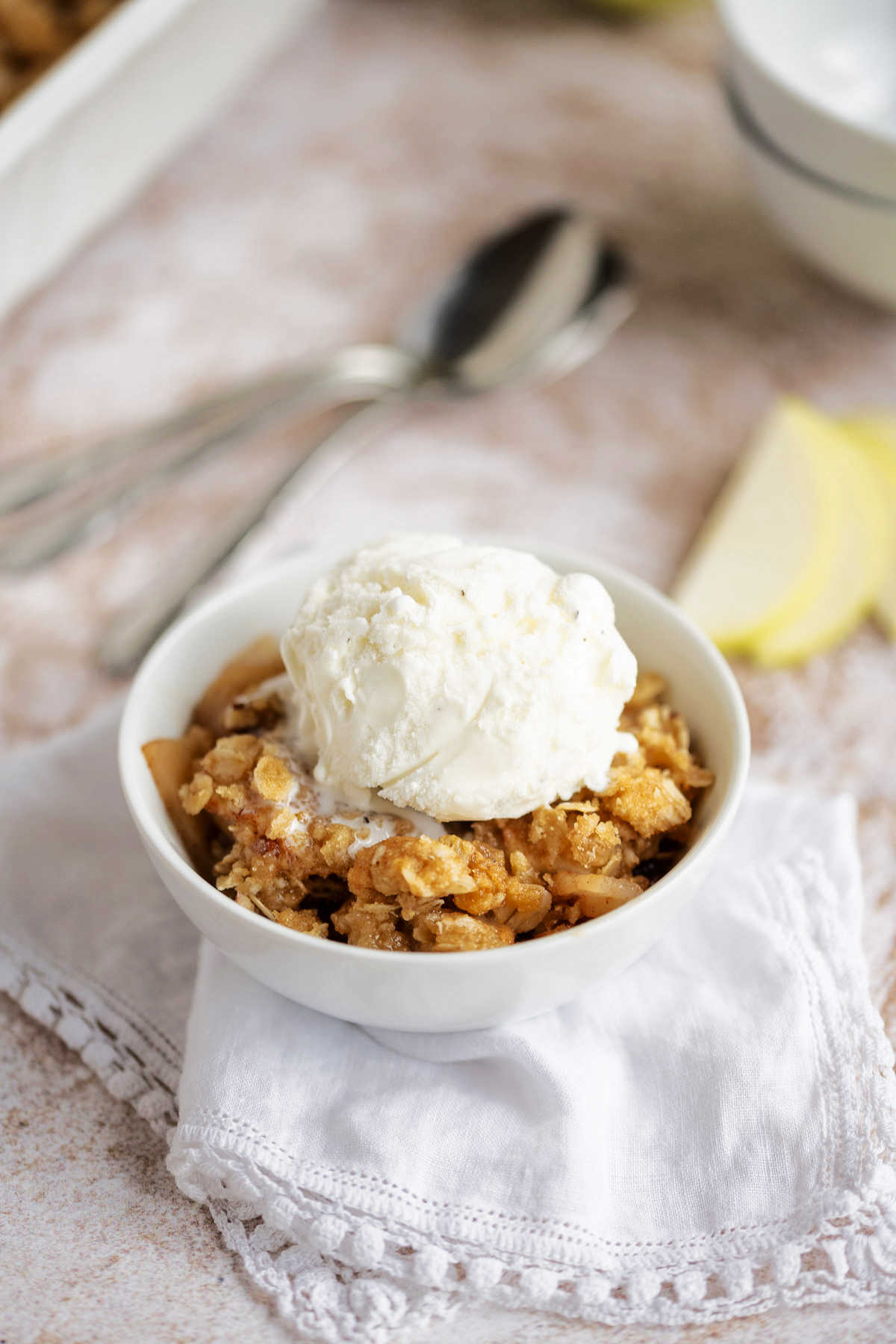 apple crisp in a white bowl with a scoop of vanilla ice cream on top sitting on a counter with apple and baking dish in the background.