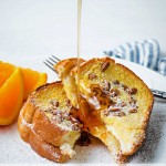 a stream of maple syrup being poured over slices of orange french toast on a white plate sitting on a table.