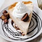 a slice of peanut butter pie on a plate with a chocolate swirl, garnished with a dollop of Cool Whip and chopped peanut butter cups sitting on a linen napkin with a gold fork.