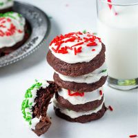 a stack of chocolate drop cookies with frosting and red sprinkles on a table with a glass of milk.