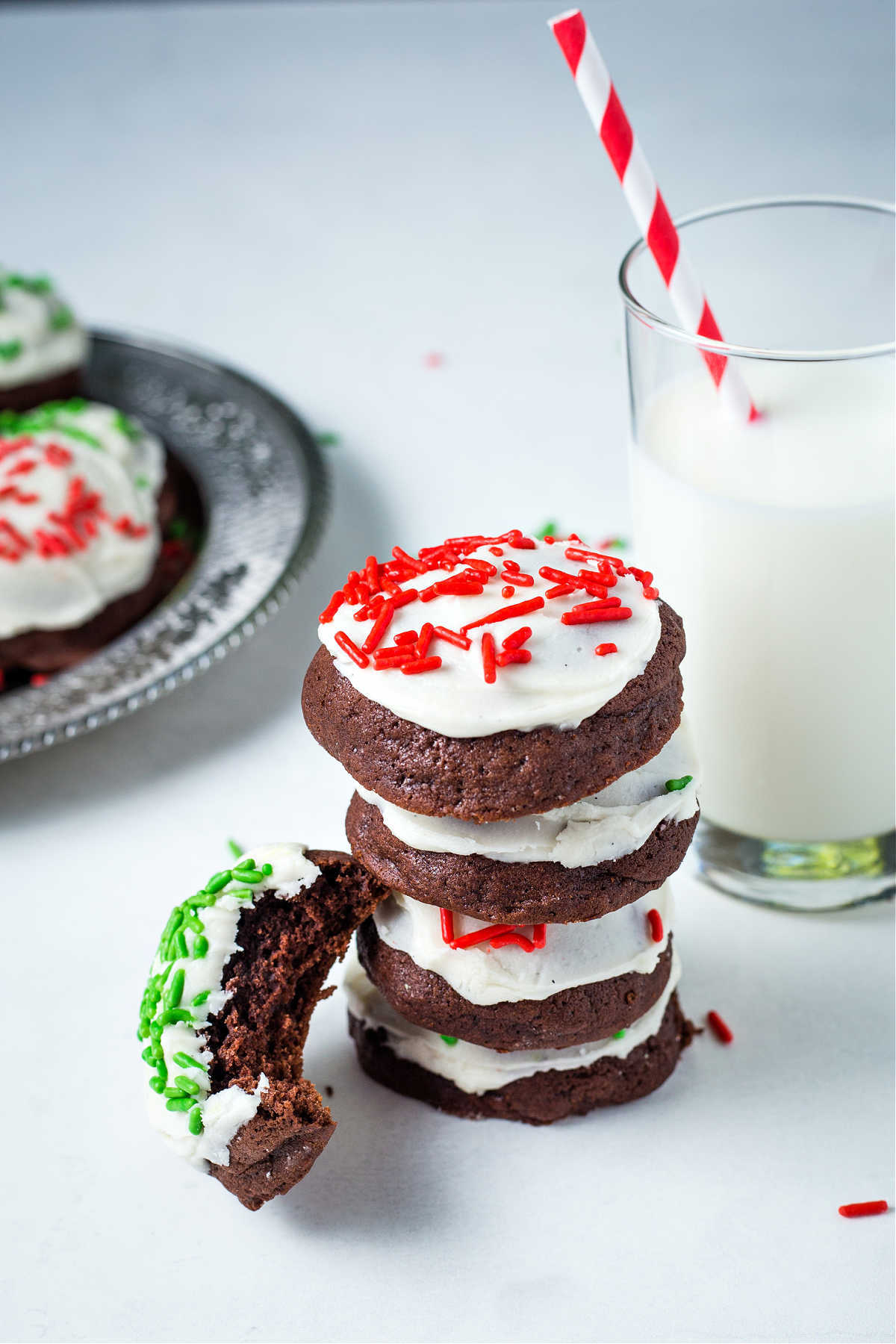 a stack of chocolate drop cookies with frosting and red sprinkles on a table with a glass of milk.