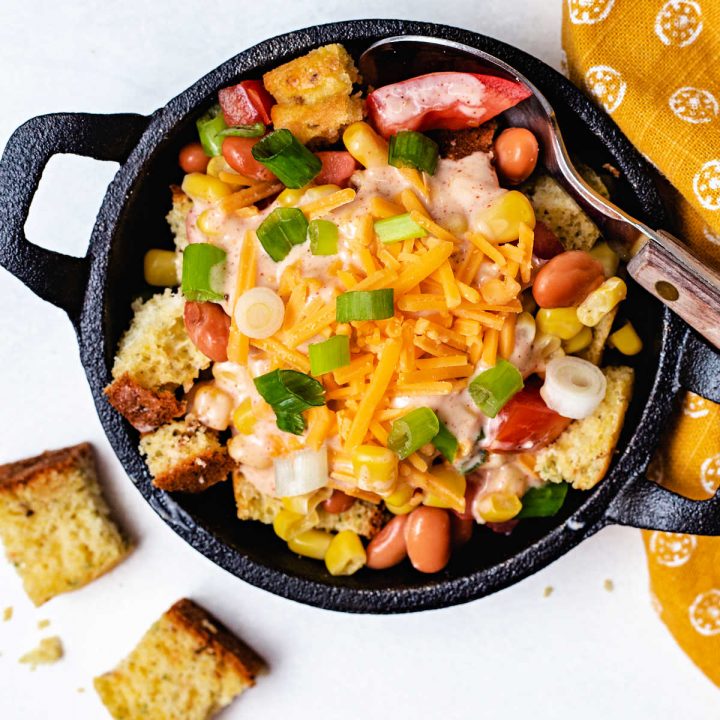 a serving of cornbread salad in a mini cast iron skilled with a spoon and yellow napkin on a table.