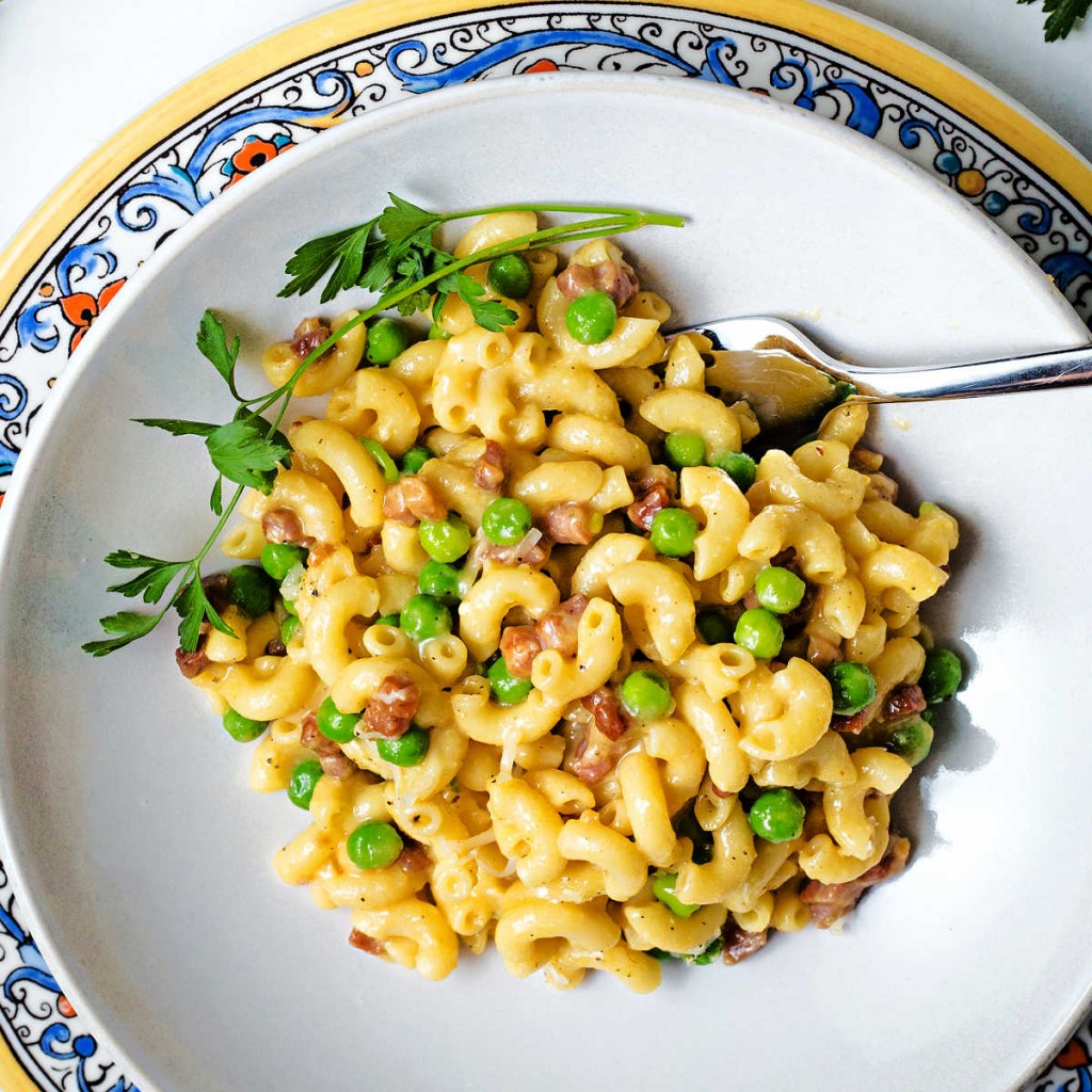 macaroni carbonara with prosciutto and green peas in a white bowl with a parsley garnish.