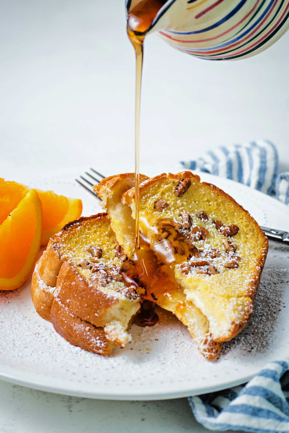 a stream of maple syrup being poured over slices of orange french toast on a white plate sitting on a table.