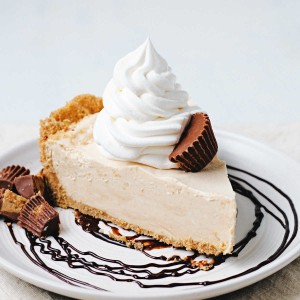 a slice of peanut butter pie on a plate with a chocolate swirl, garnished with a dollop of Cool Whip and chopped peanut butter cups sitting on a kitchen counter.