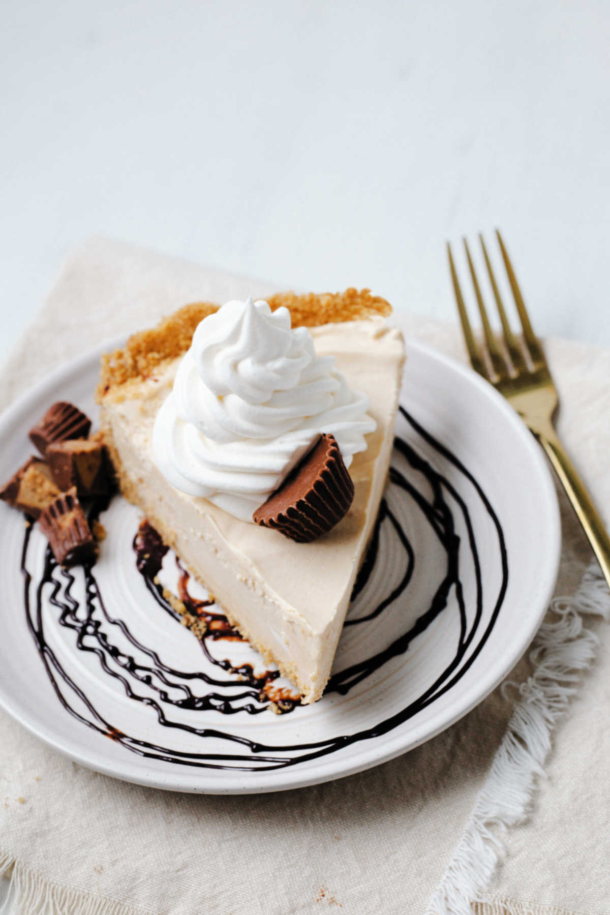 a slice of peanut butter pie on a plate with a chocolate swirl, garnished with a dollop of Cool Whip and chopped peanut butter cups sitting on a linen napkin with a gold fork.