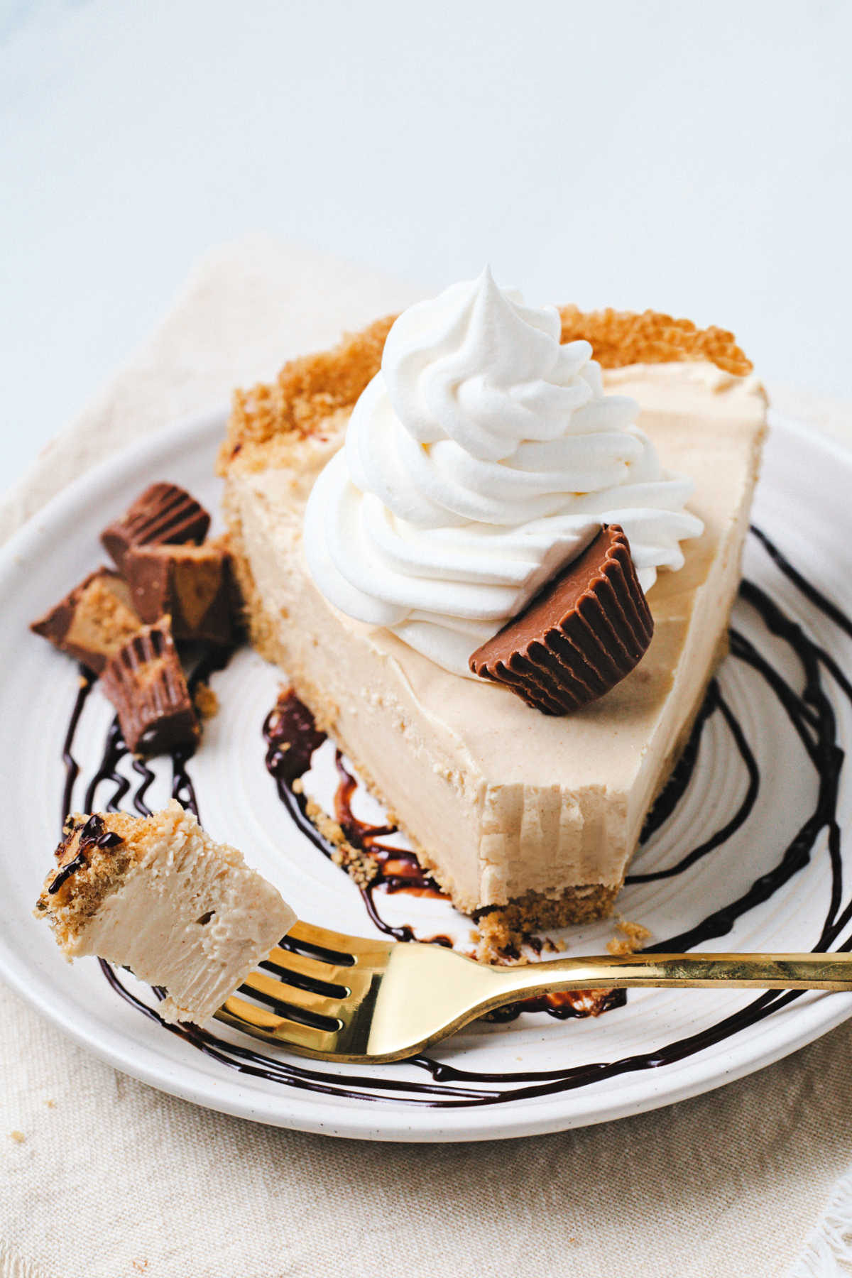 a bite of peanut butter pie on a gold fork beside a slice on a plate with a chocolate swirl, garnished with a dollop of Cool Whip and chopped peanut butter cups sitting on a linen napkin.