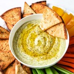 a bow of white bean hummus garnished with lemon pepper seasoning and olive oil on a platter with pita chips and veggie sticks.