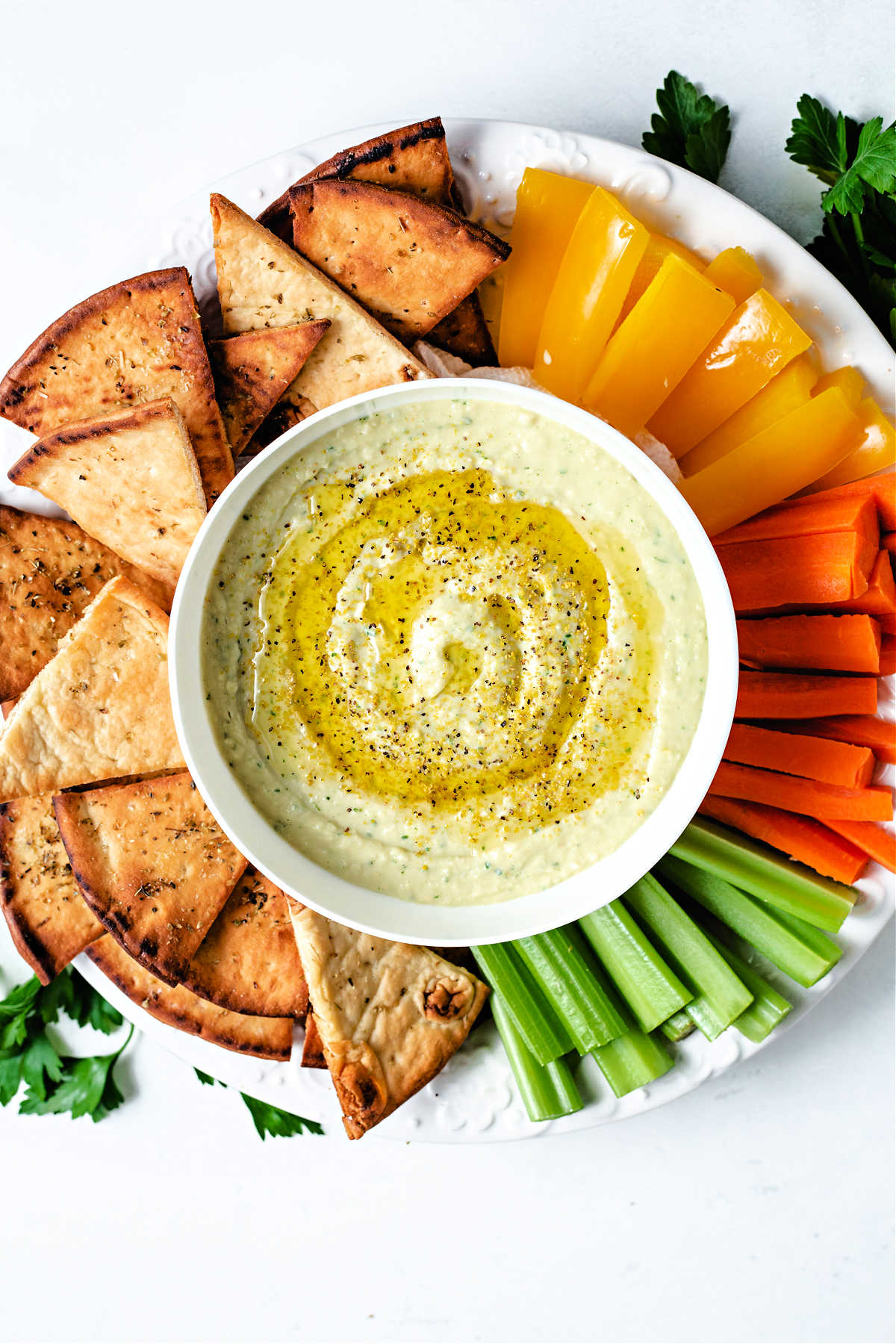 a bow of white bean hummus garnished with lemon pepper seasoning and olive oil on a platter with pita chips and veggie sticks.