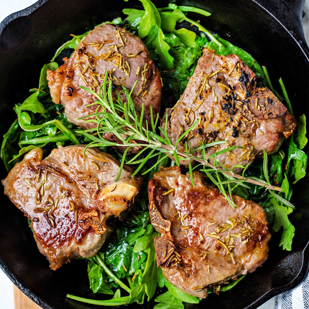 broiled lamb chops on a bed of arugula in a cast iron pan and garnished with a sprig of rosemary.