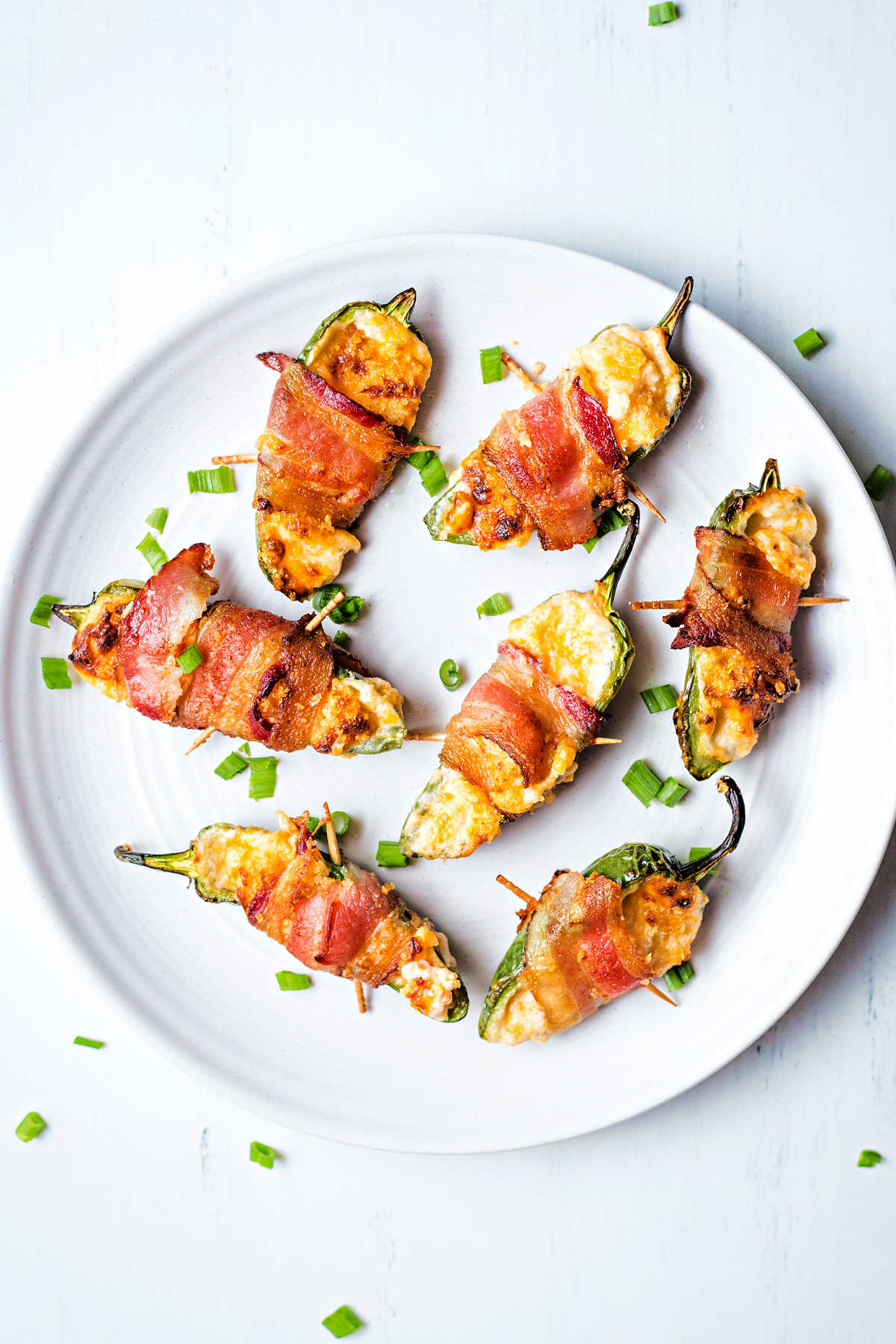 bacon wrapped jalapeno poppers on a white plate garnished with sliced green onion tops on a table.