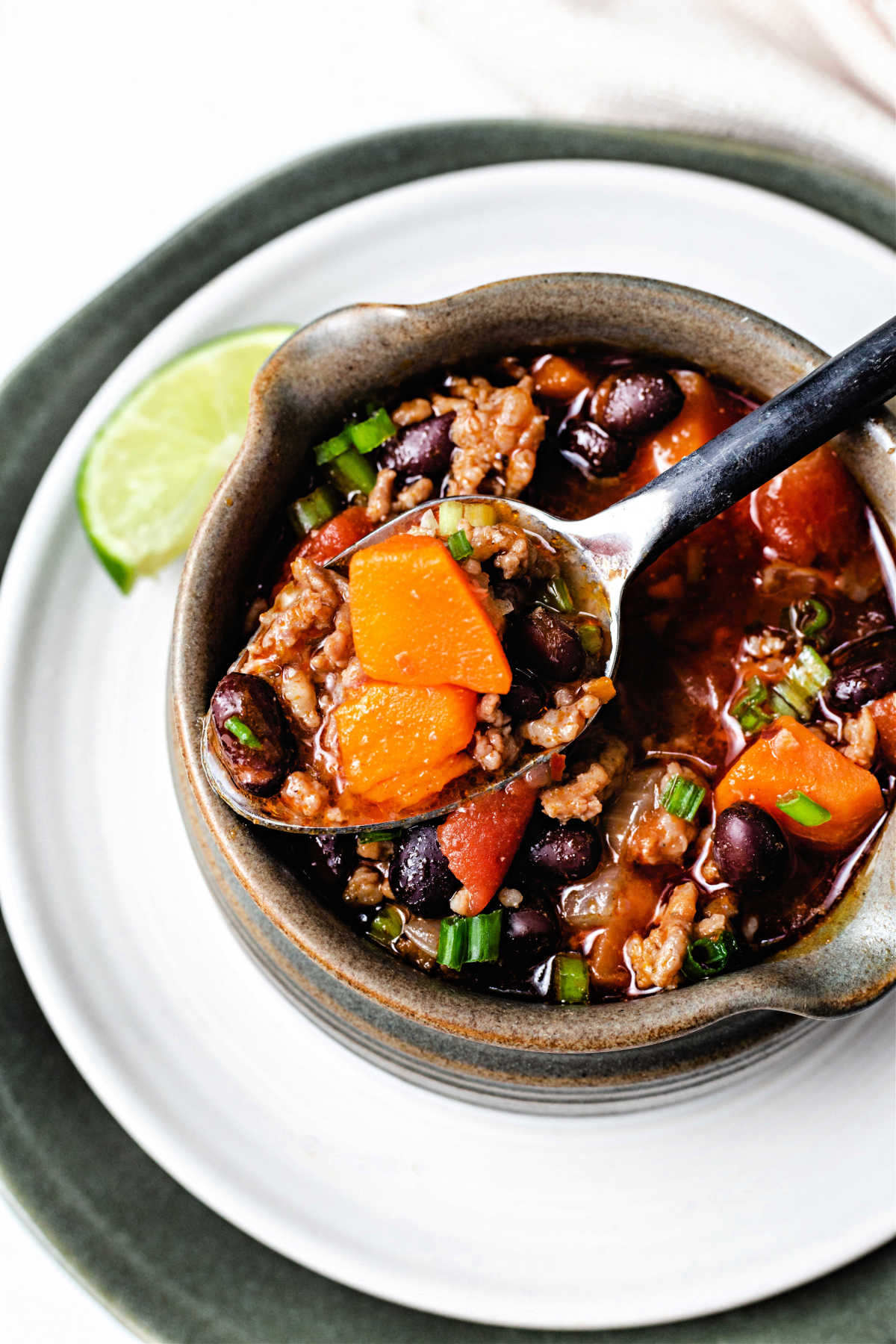 a spoonful of black bean sweet potato chili lifting out of a crock sitting on a white plate with a lime wedge.