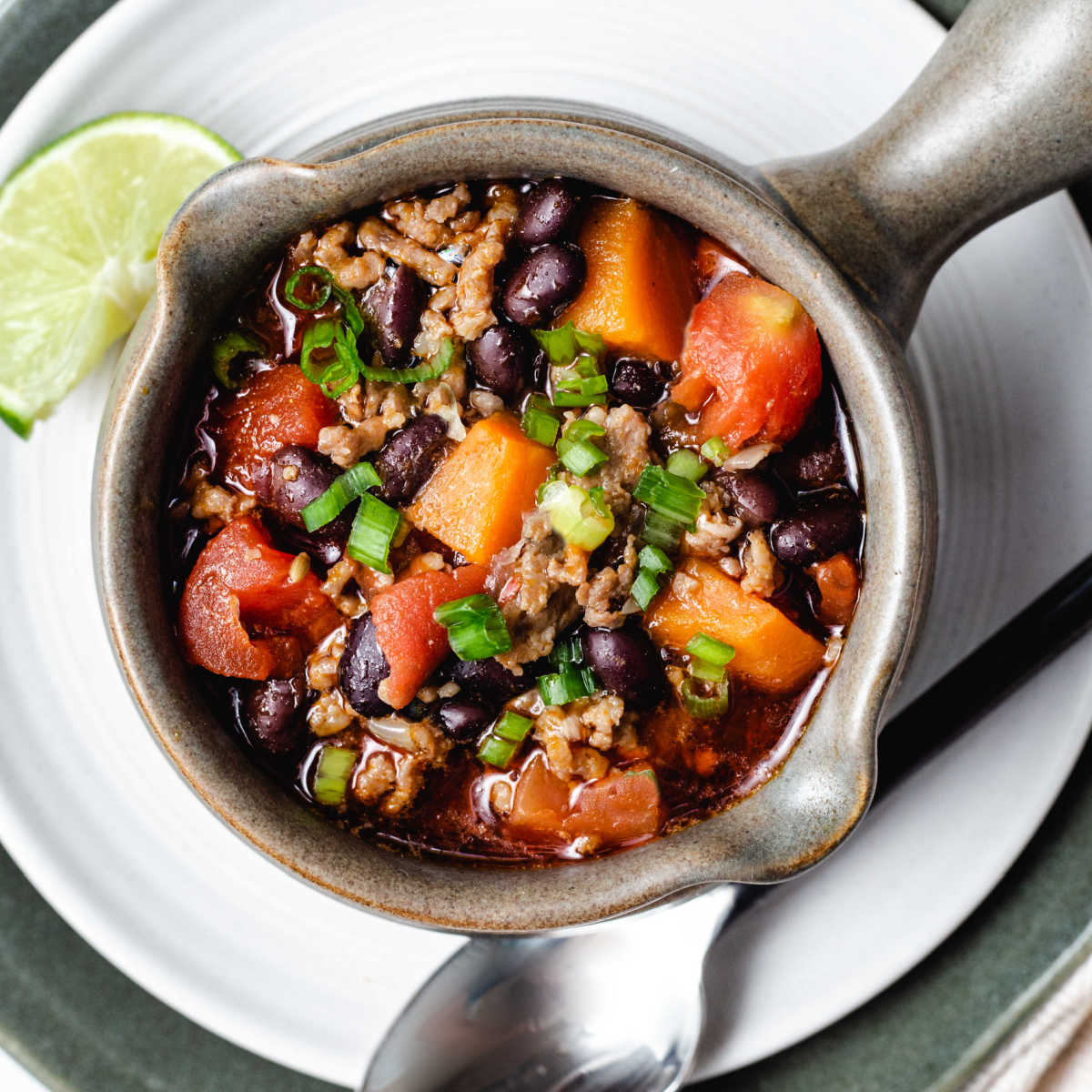 top down view of a soup mug filled with black bean and sweet potato chili sitting on a white plate with a spoon laying to the side.